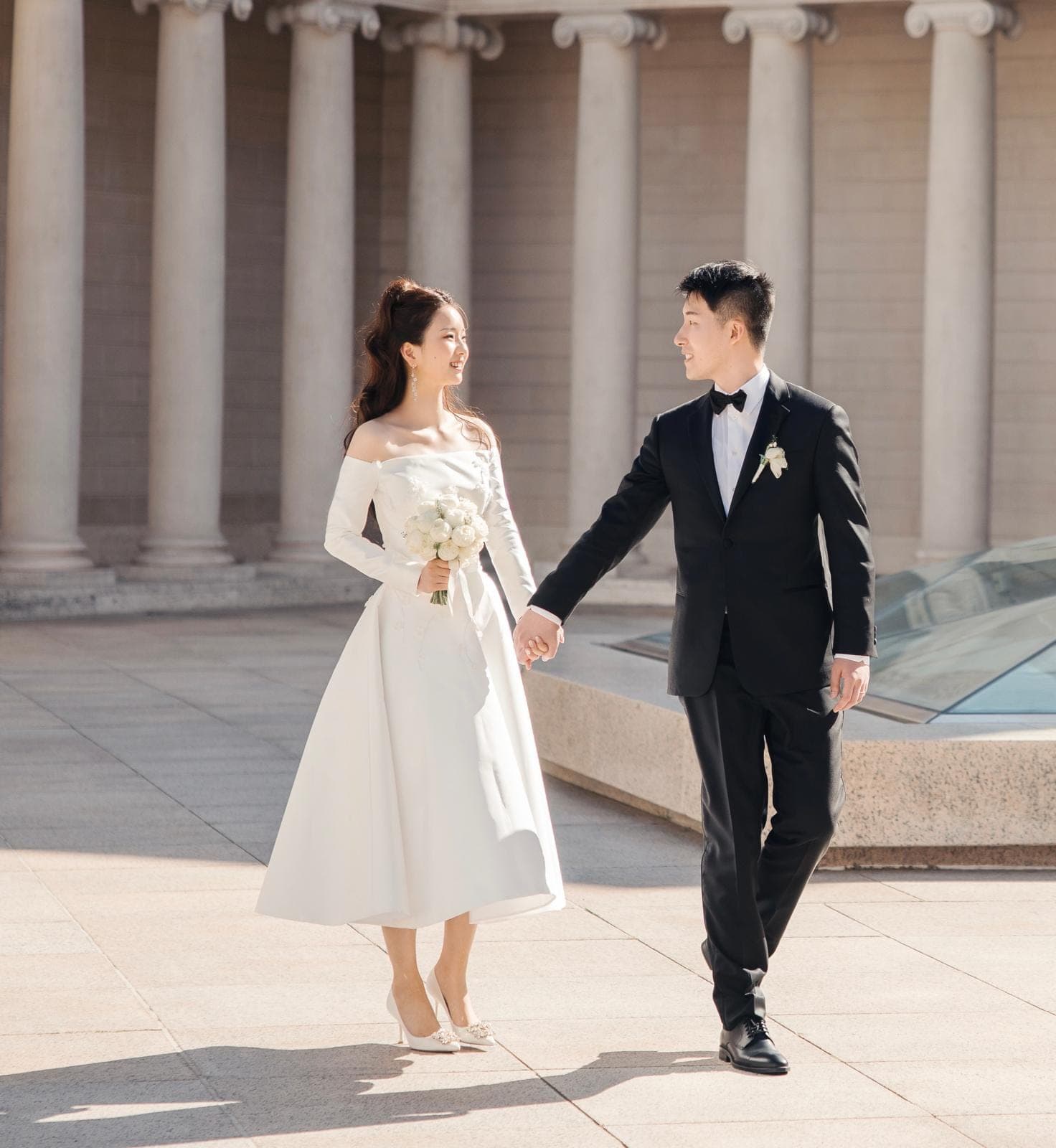 Bride and groom holding hands in front of a colonnade, she in an elegant OT Haute Couture dress