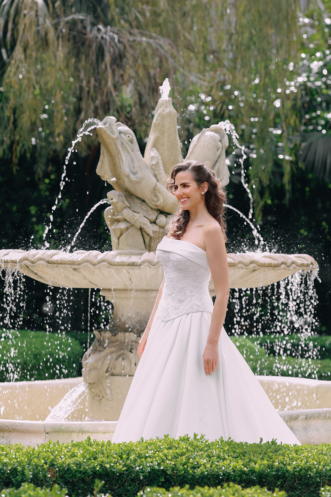 Bride wearing a luxurious beaded floral corset wedding gown with satin ballgown skirt standing in front of a garden fountain.