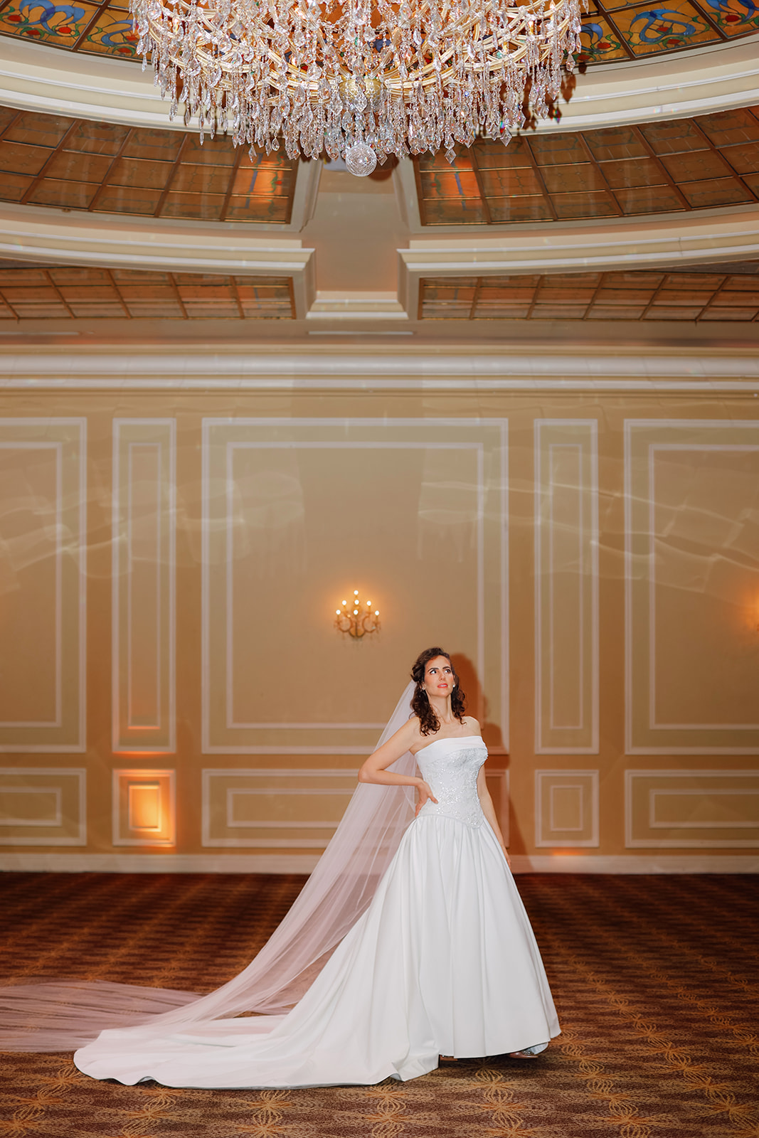 Bride wearing a luxurious beaded floral corset wedding gown with cathedral veil standing in a grand ballroom under a chandelier.